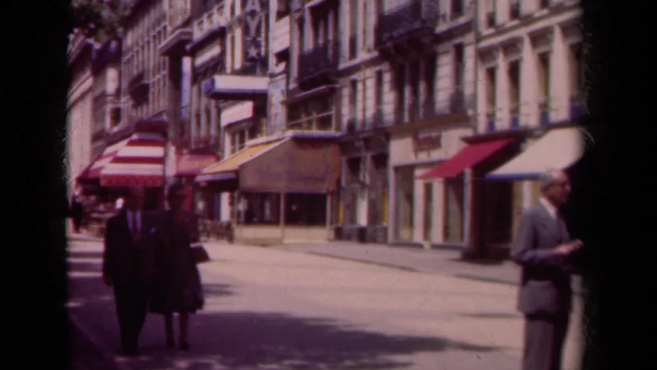 In stated 1939 Paris footage, a formally dressed couple walks toward the camera on a commercial street with awnings and apartment facades.