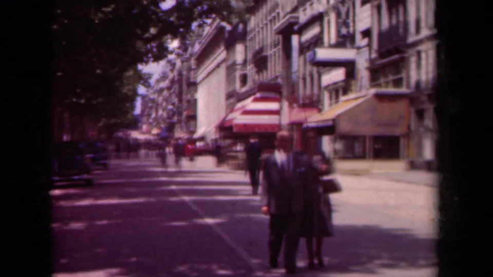 A suited man and woman with a handbag walk along the stated Paris street as awnings, trees, parked cars, and pedestrians recede behind them.