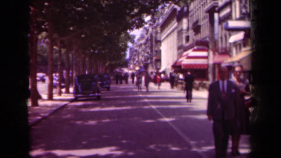 1939 Paris street view with pedestrians in period clothing, parked cars, shaded trees, storefront awnings, and a suited man partly foregrounded.