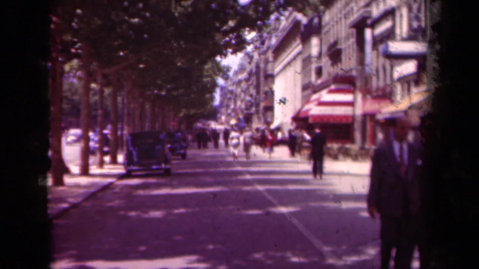 1939 Paris boulevard scene with pedestrians walking under trees, vintage cars along the curb, storefront awnings, and a cropped suited figure at right.