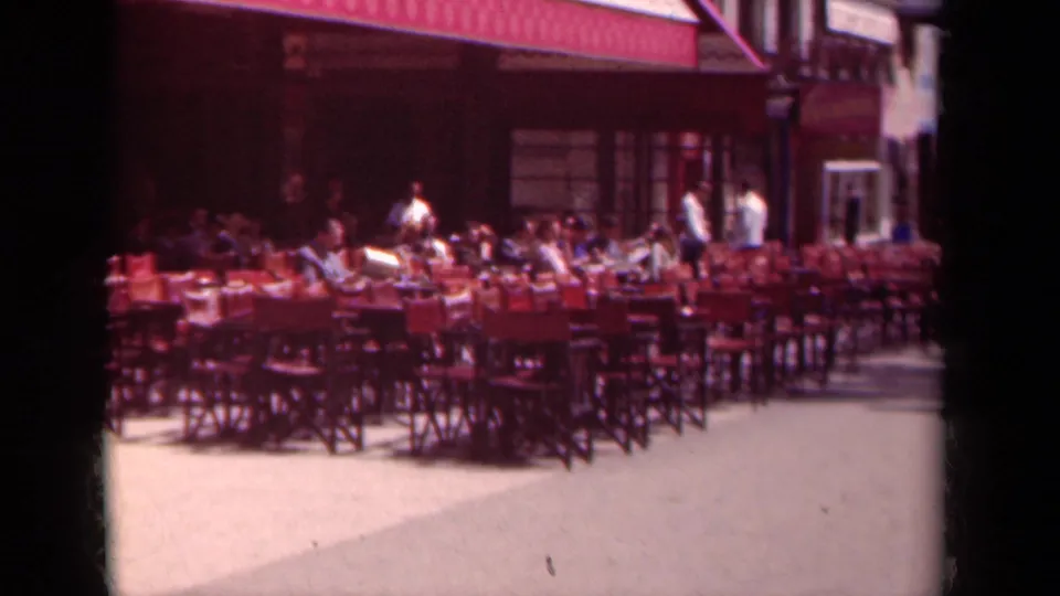 1939 Paris outdoor cafe patio with red chairs, small tables, seated patrons, staff or passersby, and a striped awning along the building front.