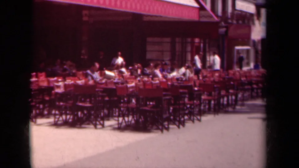 1939 Paris cafe patio showing red chairs and tables, several seated patrons, two standing figures, sidewalk space, and a striped awning across the storefront.