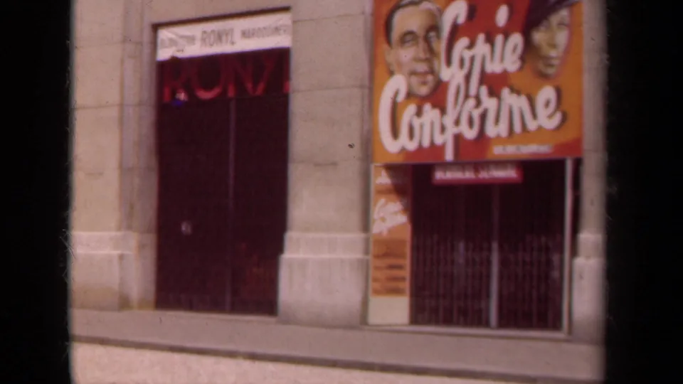 1939 Paris home-movie frame showing a red entrance, partial RONYL sign, orange Copie Conforme poster, and closed metal grille.