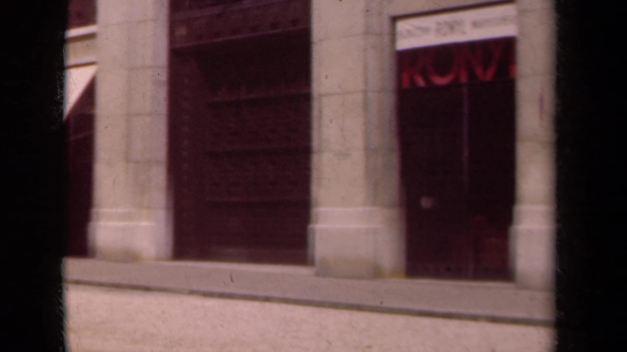 1939 Paris street view showing pale stone shopfronts, awnings, and dark entrances from a handheld 8mm perspective.