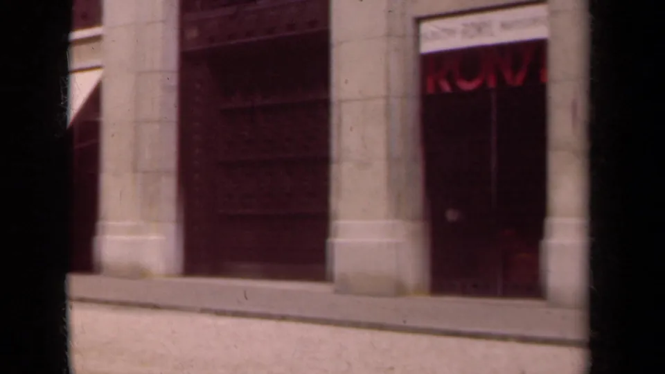 1939 Paris street view showing pale stone shopfronts, awnings, and dark entrances from a handheld 8mm perspective.