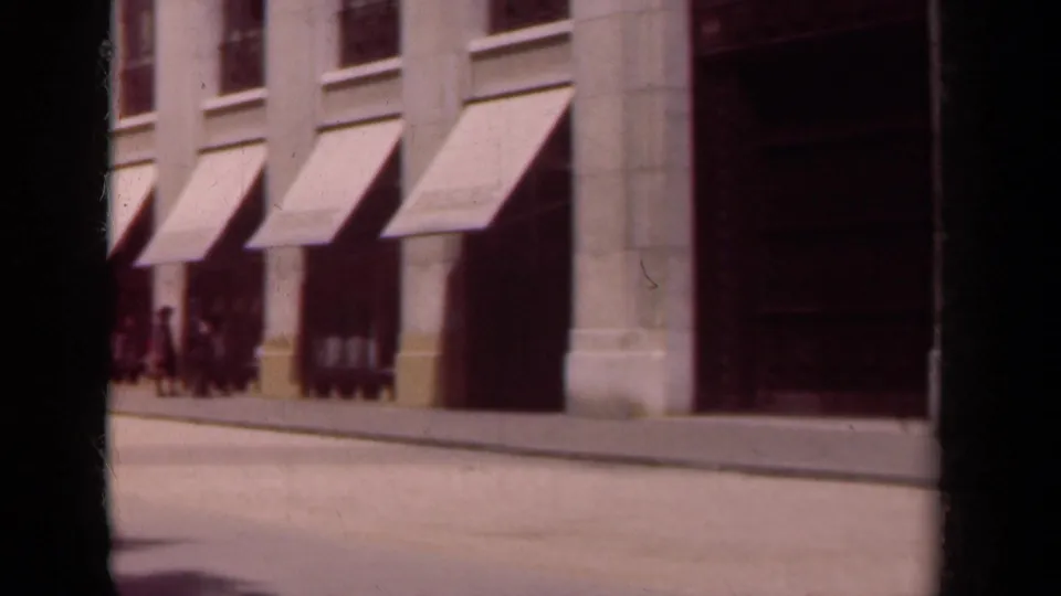 1939 Paris pedestrians gather near awning-covered storefronts as the camera looks along the sidewalk.