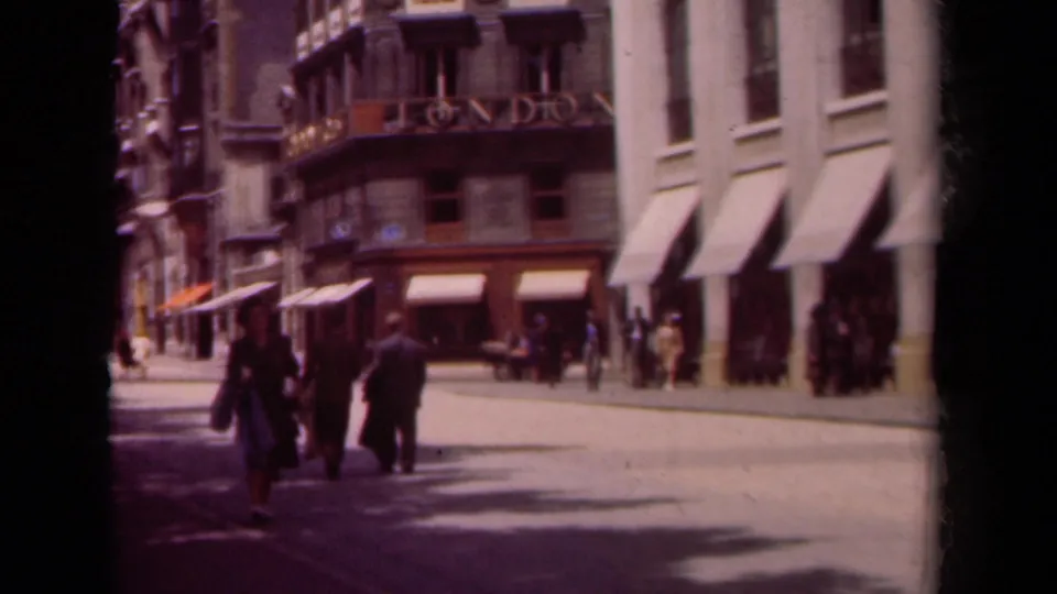 1939 Paris street corner with pedestrians crossing, shop awnings, upper balconies, and bright pavement shadows.