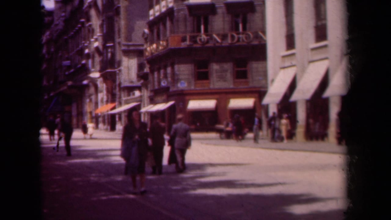 Pedestrians stand and walk on a Paris boulevard near a corner building with awnings and signage.