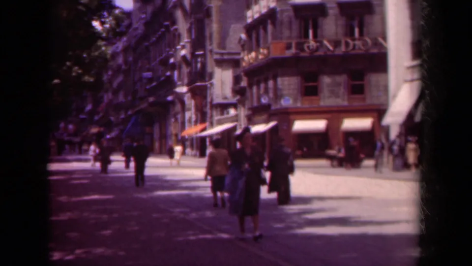 1939 Paris boulevard view with pedestrians moving past shops, trees, street lamps, and a building carrying visible signage.