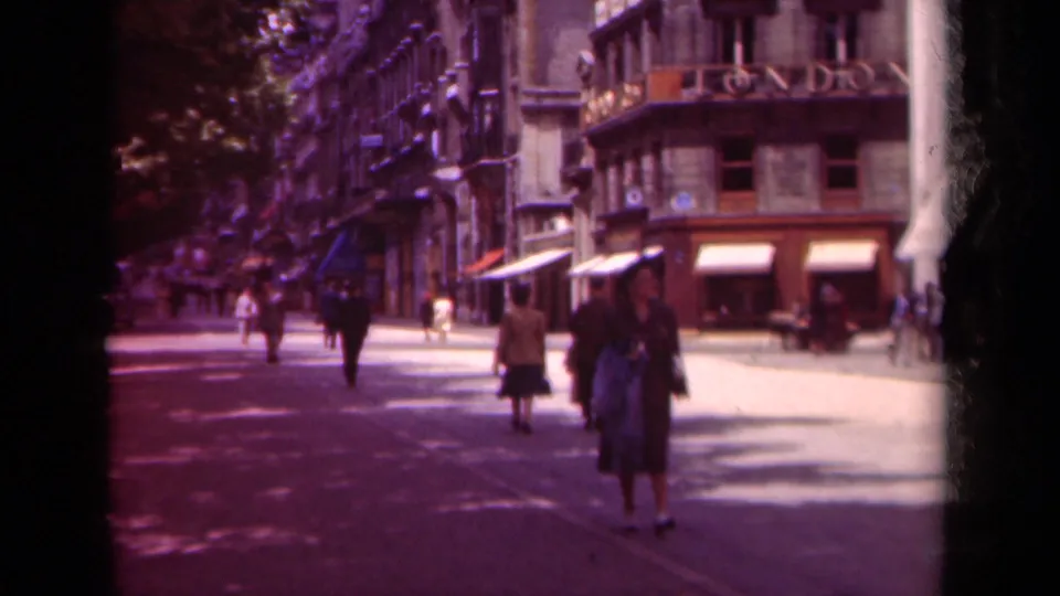 1939 Paris street view with a woman approaching the camera, other pedestrians nearby, and shop awnings along a tree-lined boulevard.
