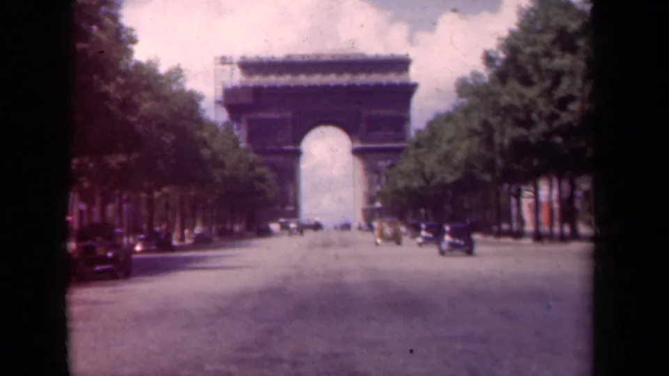 1939 Paris frame with the Arc de Triomphe in the distance, cars moving along the Champs-Elysees, and trees on both sides.
