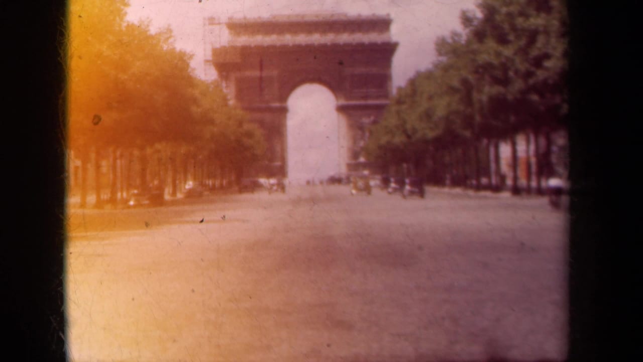 1939 Paris home-movie frame looking along a broad avenue toward the Arc de Triomphe, with trees and faint street traffic visible.