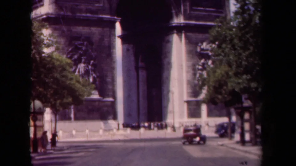 Street-level 1939 Paris frame near the Arc de Triomphe, showing a classic car, pedestrians, trees, and part of the monument.