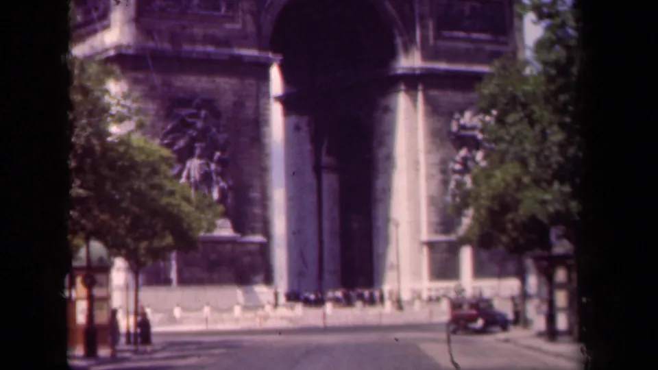 1939 Paris home-movie frame showing the Arc de Triomphe at street level, with cars, pedestrians, trees, and dark film borders.