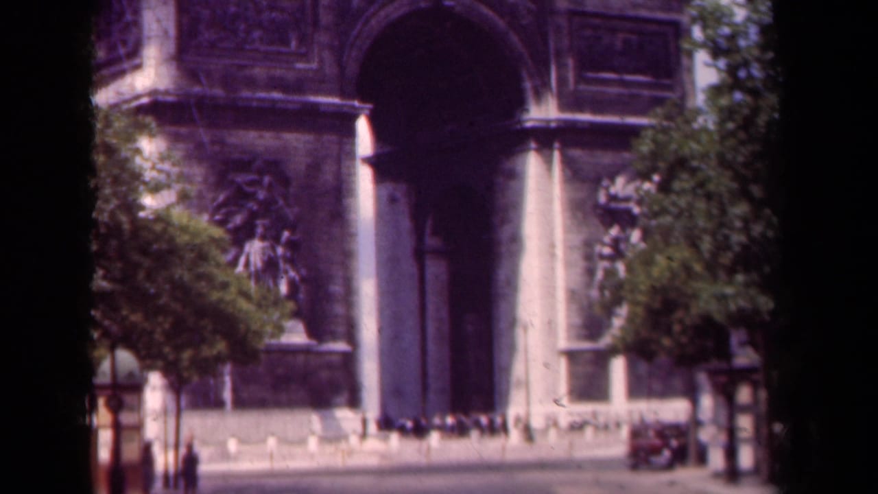 1939 8mm view of the Arc de Triomphe in Paris, framed by trees with small pedestrians and a vintage car near the monument.