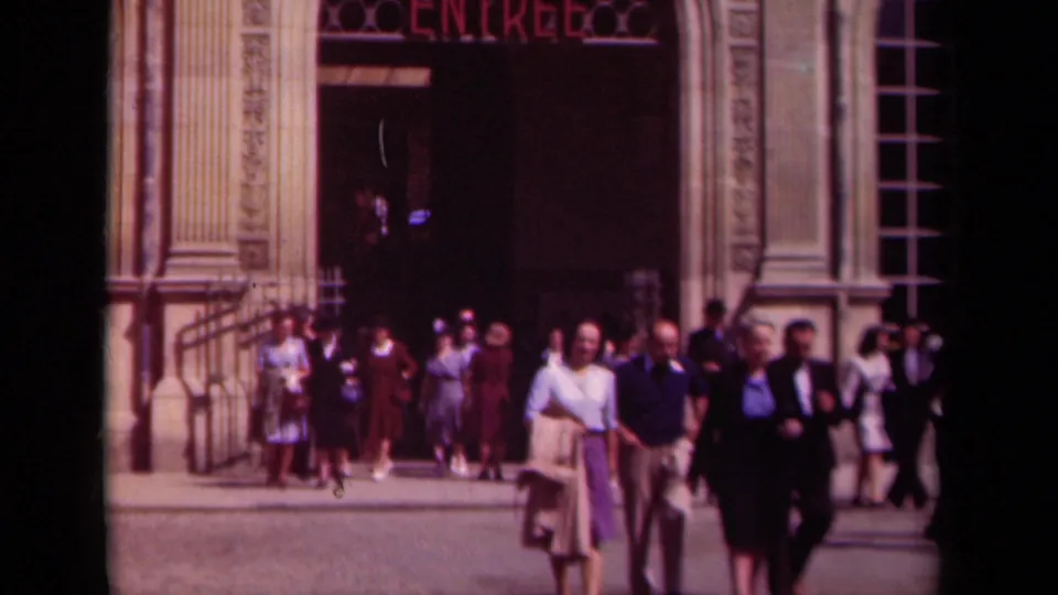 People in 1930s clothing walk near a Paris doorway marked ENTREE, with faces and architectural columns visible.