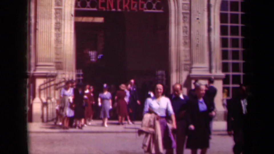 A crowd exits or passes by a Paris entrance, with period dresses, suits, railings, and a tall window visible.
