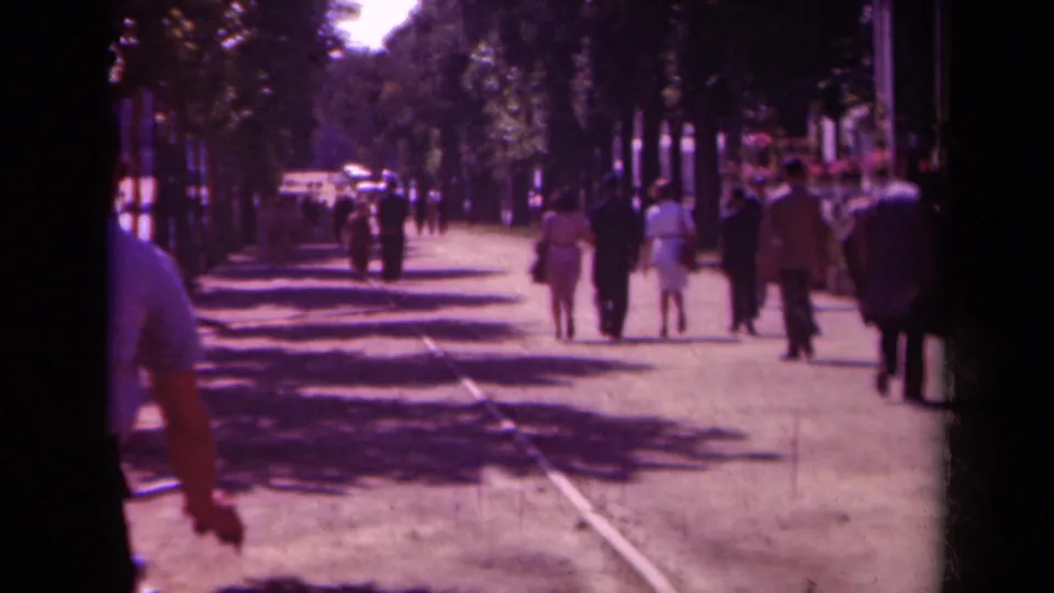 1939 Paris home-movie frame with a partial cyclist in the foreground and pedestrians walking away on a tree-lined street.