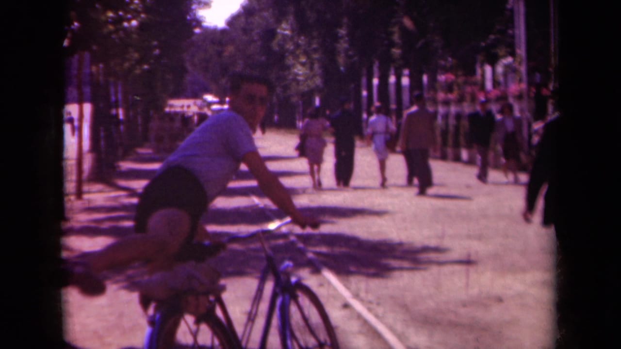 Man dismounting a bicycle beside a busy tree-lined walkway with pedestrians in the distance.