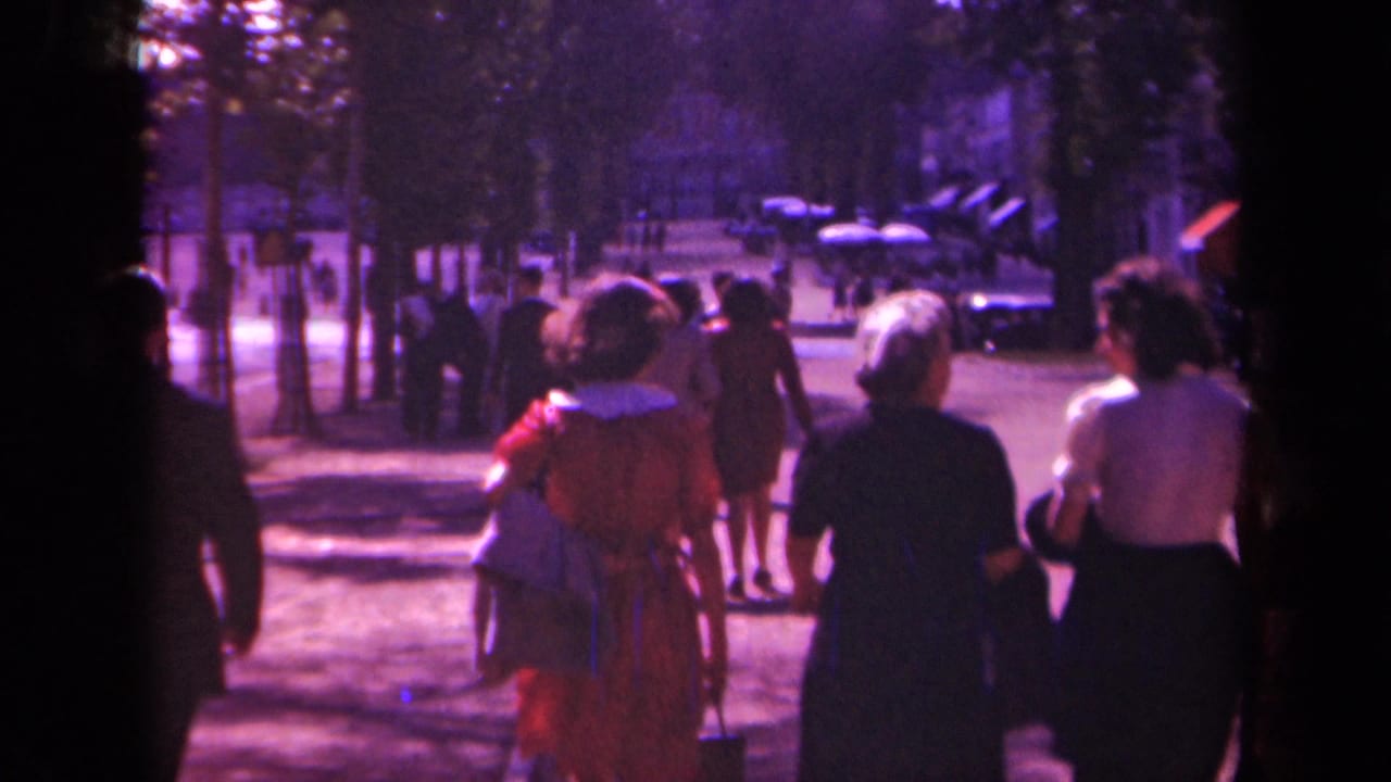 1939 Paris street scene with adults in period clothing walking along a tree-lined boulevard, seen through soft faded 8mm color.