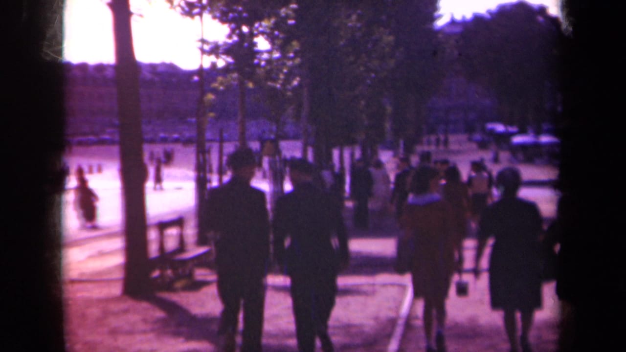 1939 Paris home-movie frame showing silhouetted pedestrians on a tree-lined public walkway beside a broad street.