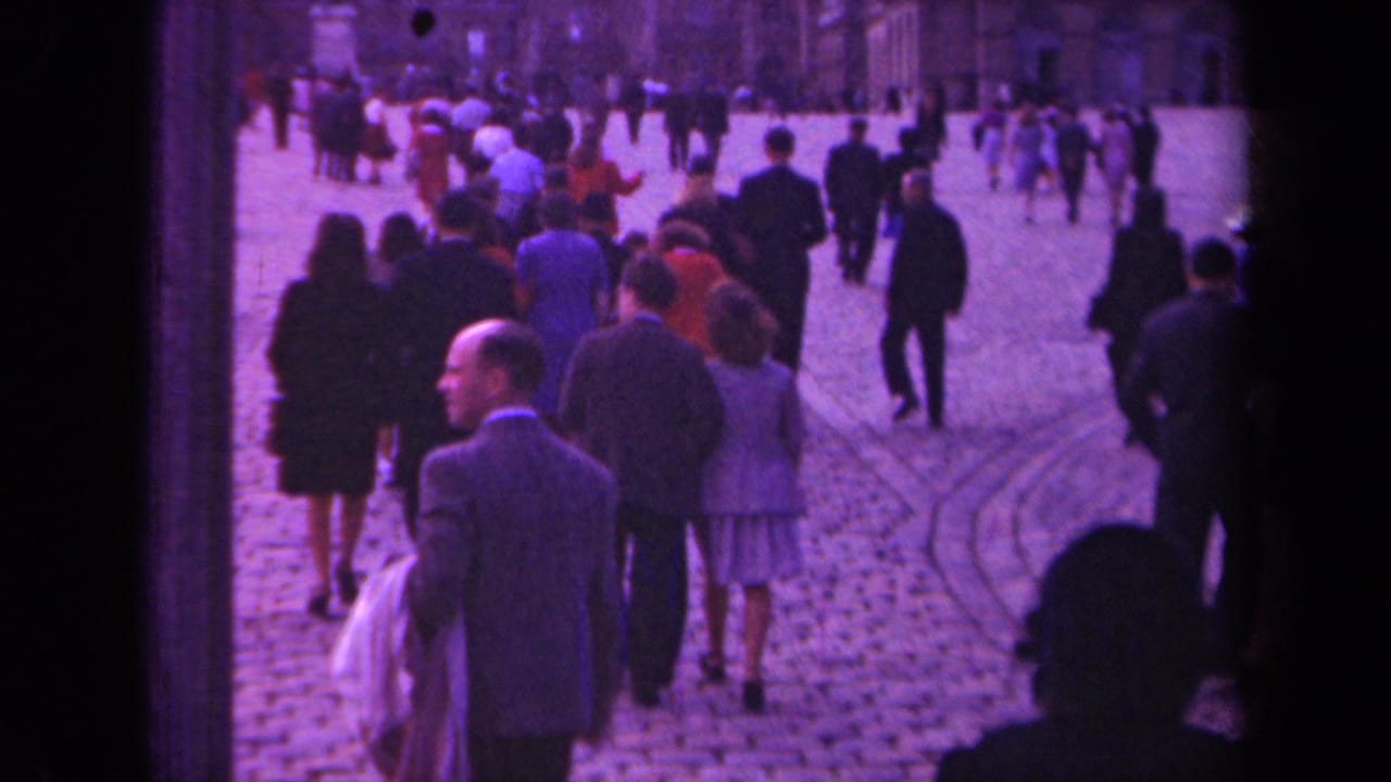Paris 1939 street footage frame with pedestrians crossing a cobblestone square and a suited man near the center foreground.