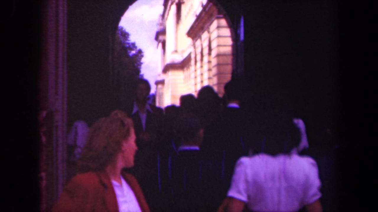 In 1939 Paris home-movie footage, tourists move through a shadowed arch toward sunlit buildings beyond.