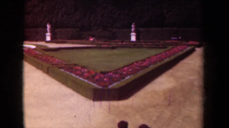 The garden view holds on a triangular planted bed bordered by flowers and hedges, with statues and a small visitor partly visible below.