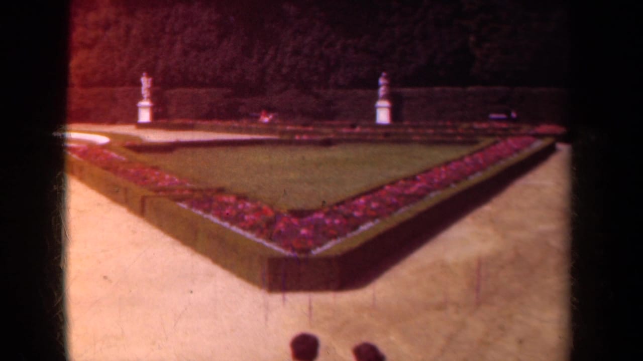 1939 Paris home-movie view of a formal garden with a triangular flowerbed, clipped hedges, gravel paths, lawn, and distant statues.