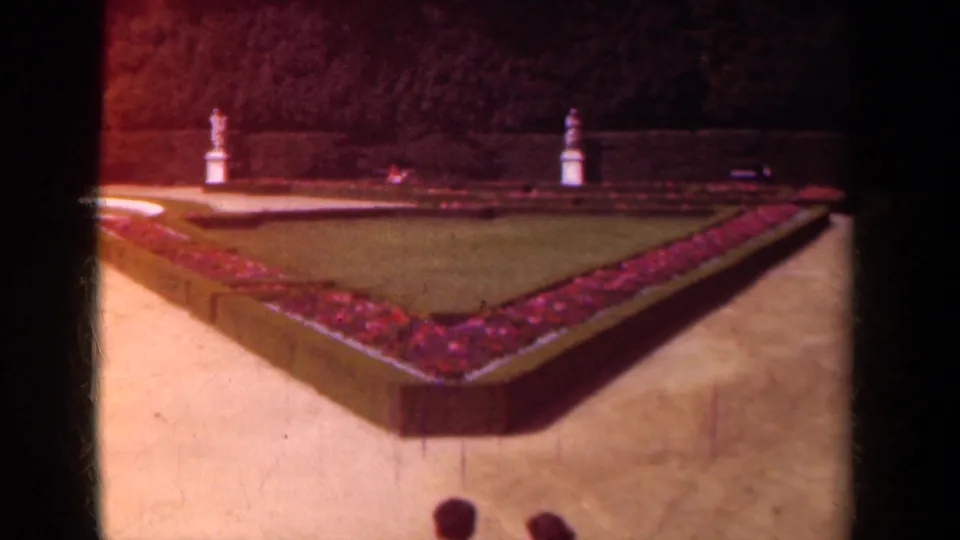 1939 Paris home-movie view of a formal garden with a triangular flowerbed, clipped hedges, gravel paths, lawn, and distant statues.