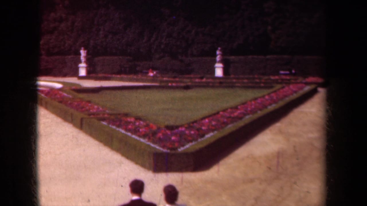 In Paris in 1939, a couple is seen from behind near a formal garden with a triangular flowerbed, clipped hedges, gravel paths, and statues.