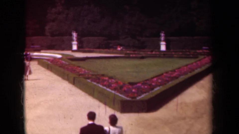 1939 Paris footage shows a couple walking away along a gravel path beside a triangular flowerbed, lawn, clipped hedges, and distant statues.