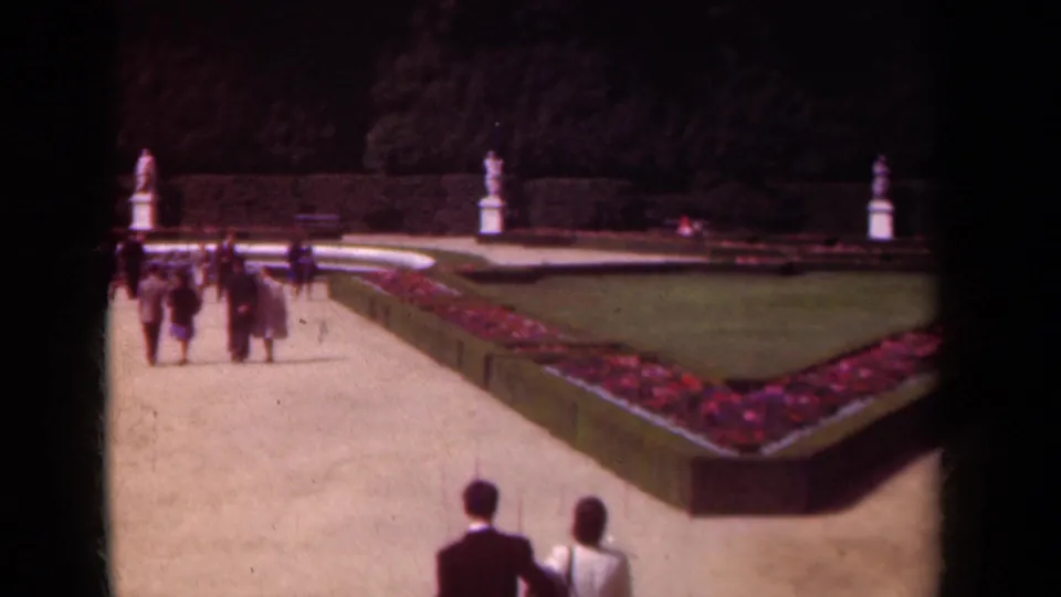 1939 Paris garden scene with multiple visitors on gravel paths around a triangular flowerbed, clipped hedges, lawn, and classical statues.
