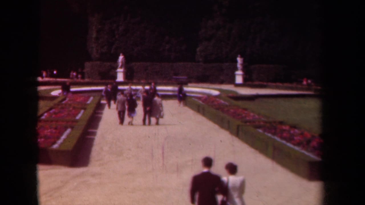 1939 Paris garden view with couples in period clothing walking toward white statues beyond clipped hedges and bright flowerbeds.