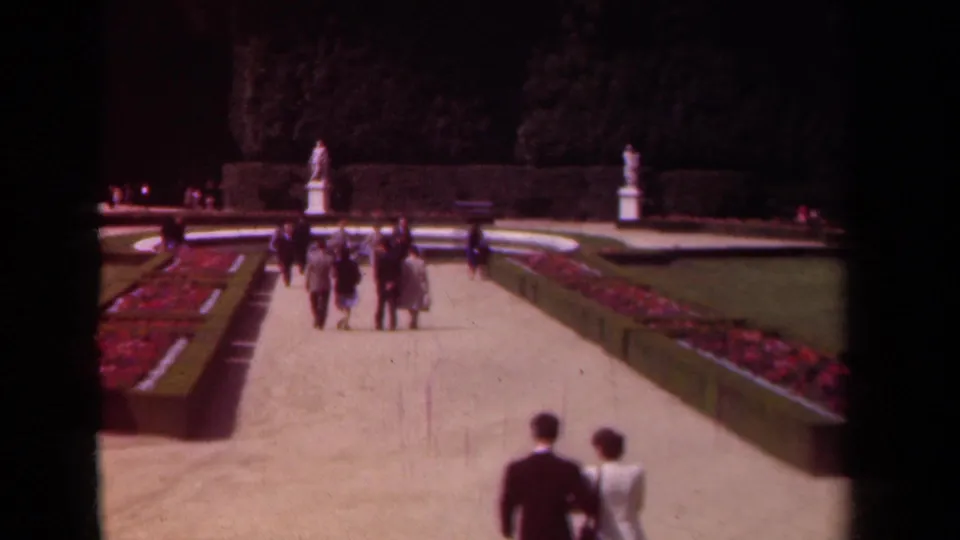 1939 Paris garden view with couples in period clothing walking toward white statues beyond clipped hedges and bright flowerbeds.