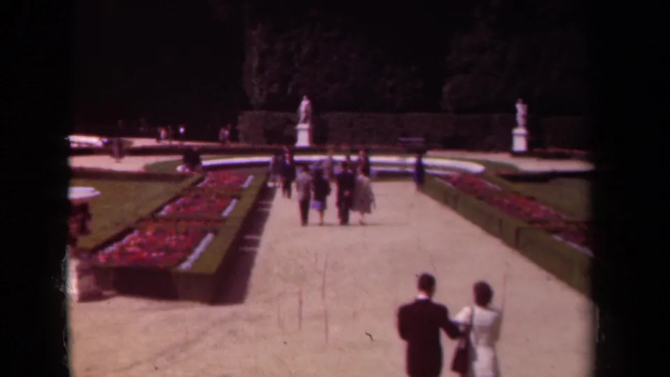 1939 Paris home-movie frame with a foreground couple and distant visitors walking past red flowerbeds, lawns, hedges and white garden statues.