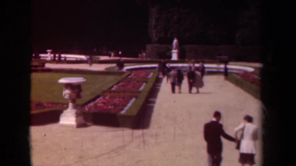 1939 Paris garden frame showing a couple at lower right, a stone urn, red flowerbeds, lawns, hedges, benches and visitors in the distance.