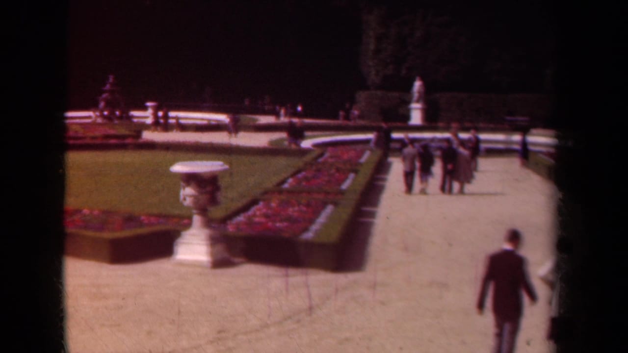 Visitors walk along a formal garden path beside flower beds, lawn, and a white urn.