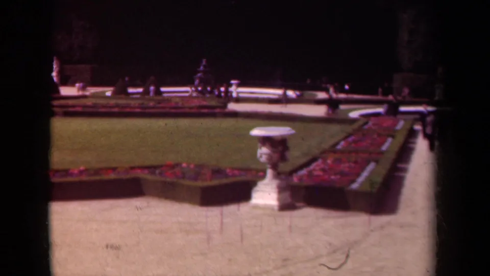 Formal Paris garden scene from 1939 with a white urn, red planting beds, gravel paths, lawn, distant visitors, and statuary.