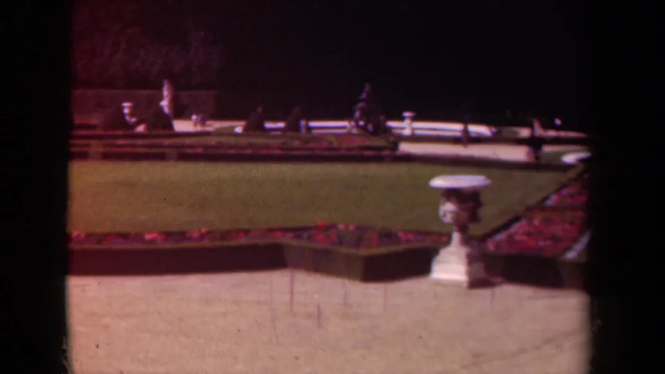 1939 Paris formal garden view with red flower beds, lawn, a white urn, distant visitors, statues, and soft 8mm color.