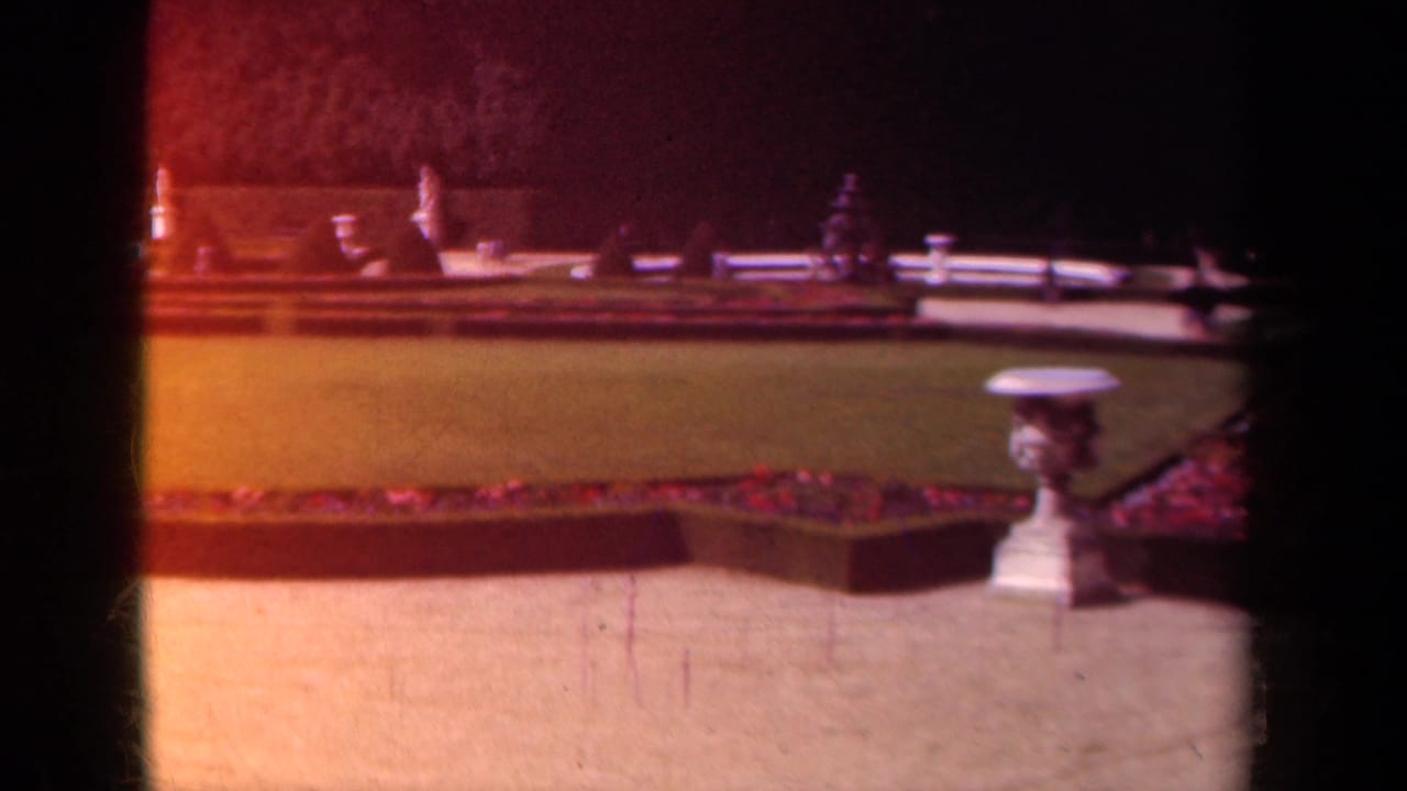 1939 Paris home-movie frame showing a formal garden lawn, red flowerbed edging, stone urn, and shadowed foreground obstruction.