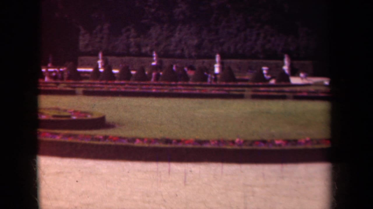 1939 Paris home-movie frame showing a formal garden with circular flower bed, clipped lawn, hedges, and a tree-lined background.