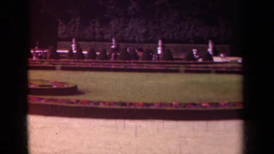 1939 Paris home-movie frame showing a formal garden with circular flower bed, clipped lawn, hedges, and a tree-lined background.