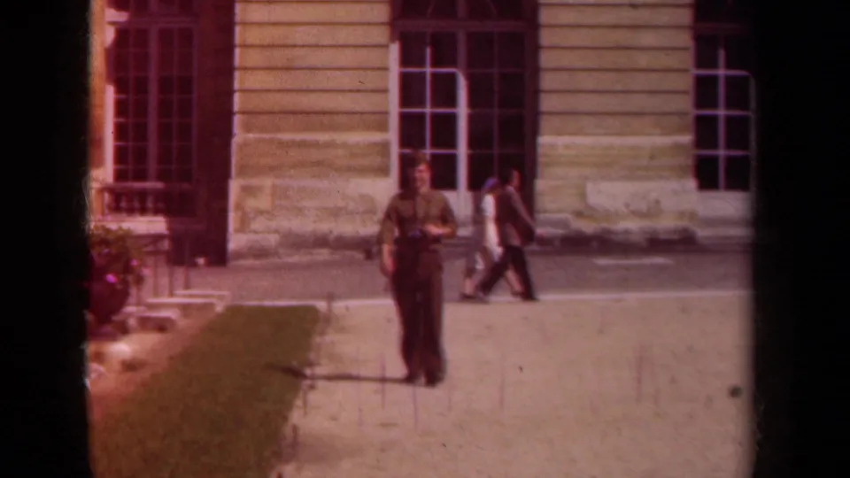 1939 Paris frame showing a uniformed man walking across formal grounds near a yellow building while another adult passes in the background.