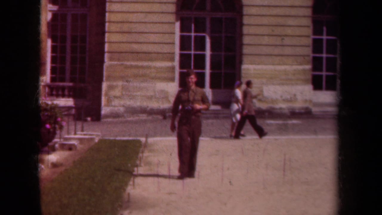 Uniformed man walking on a paved path in front of a stone building with pedestrians behind him.