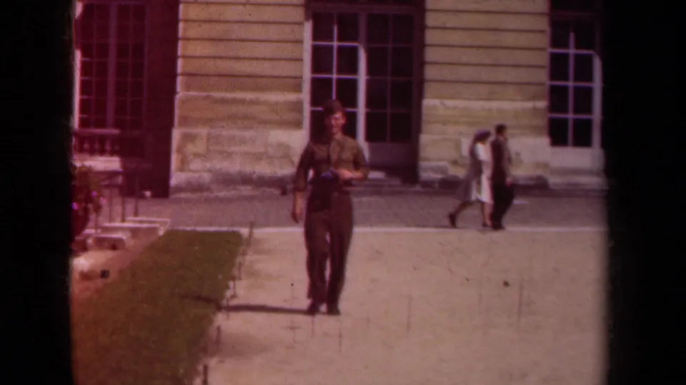 The man in uniform, wearing a camera at his chest, moves forward across the paved area in front of a Paris stone facade.