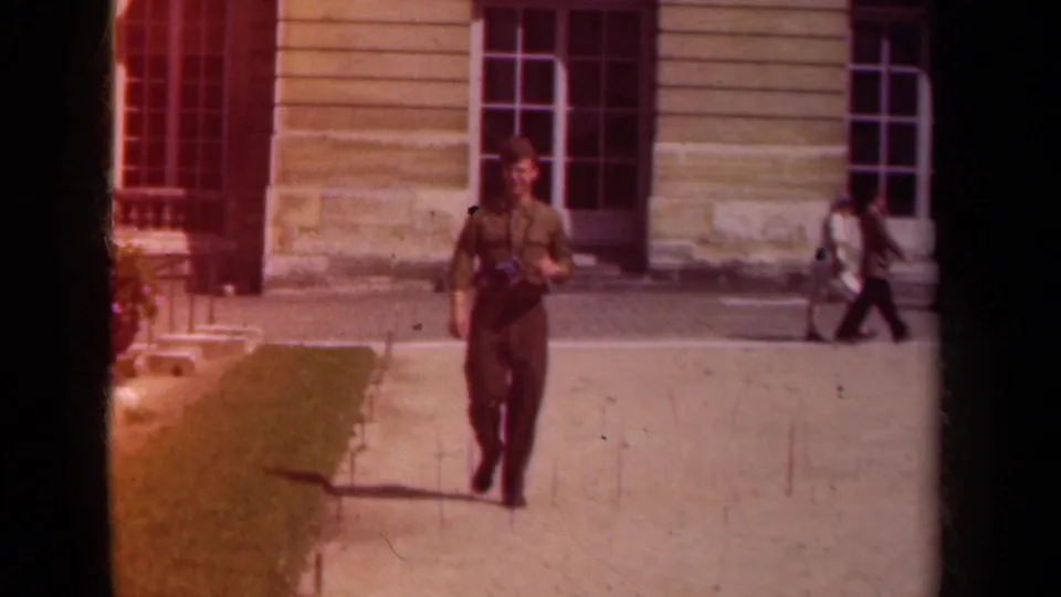 A uniformed adult approaches the camera in the Paris setting, with a camera on his chest and pedestrians crossing behind him.
