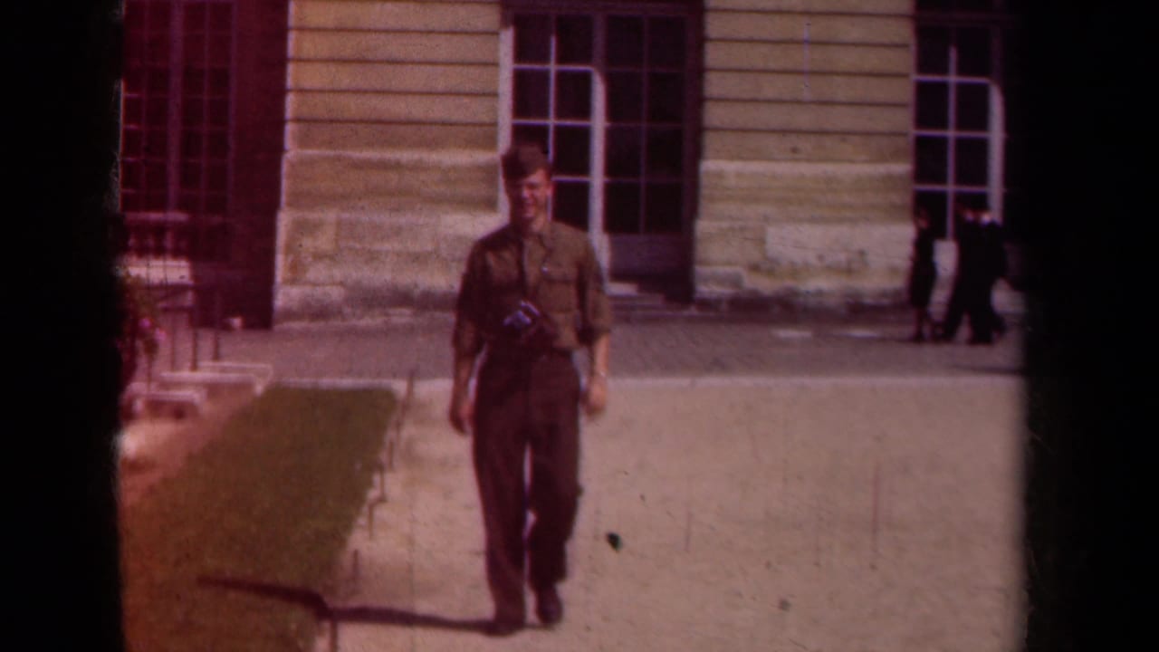 Uniformed soldier walking toward the camera in a courtyard near a stone building.
