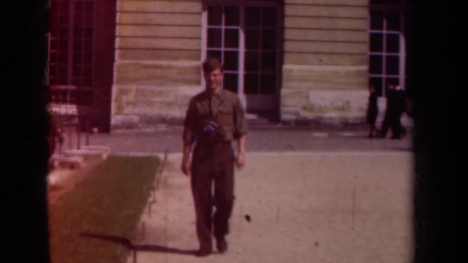 In Paris, France, a uniformed soldier walks toward the camera with a camera hanging from his neck in a sunlit courtyard.