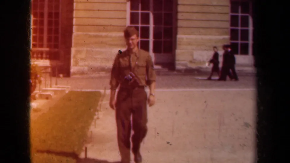The soldier continues toward the camera in a Paris courtyard, smiling faintly with a camera strap across his uniform.
